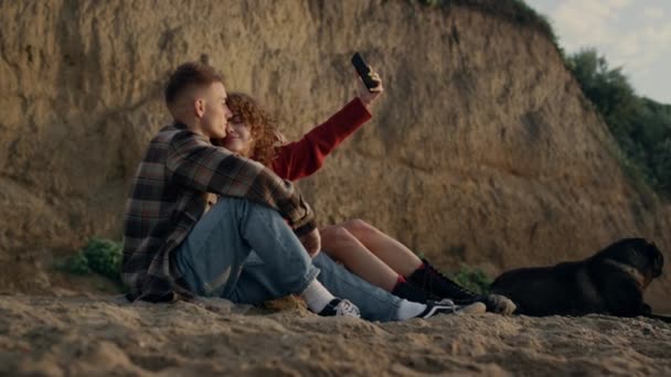 Hellyttävä pari ottaa selfie valokuvan älypuhelimella auringonlaskun rannalla. Onnellinen nainen ja mies katsomassa kuvia matkapuhelin. Nuoret rakastavaiset, joilla on koira hiekkarannalla. Iloiset hipsterit halailevat yhdessä. - Materiaali, video