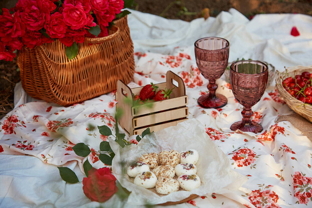 Γυναικείο ροζ καλοκαιρινό πικ-νικ με τραπεζομάντηλο, ζαχαρωτά, φράουλες, κεράσι, μπιζέλια και ποτήρια κρασί ανάμεσα σε ροζ τριαντάφυλλα στο πάρκο. Ρομαντικό βράδυ, το ταξίδι και την εκτίμηση της στιγμής - Φωτογραφία, εικόνα