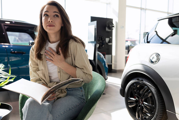 egy fiatal nő egy autókereskedésben tanulmányozza a katalógust egy sor új autóval.. - Fotó, kép