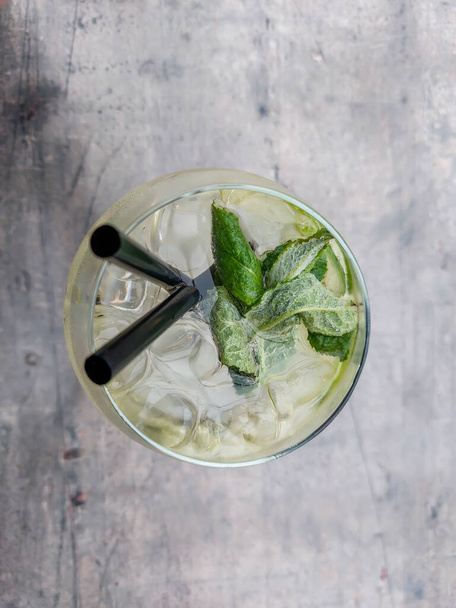 Coctail Mojito with green mint leaves, white rum, lime, lemon, soda and ice cubes on a table in an outdoor setting. Pieces of lime and mint in big glass - Foto, imagen