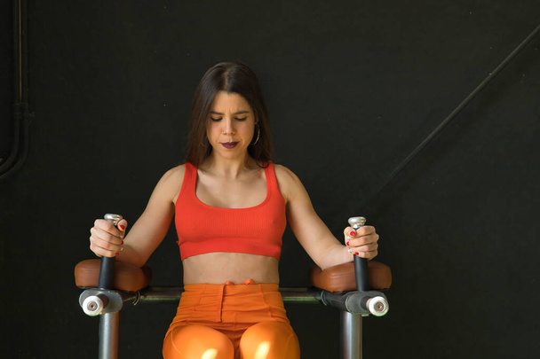 Νεαρή όμορφη γυναίκα ακουμπά στο κοιλιακό μηχάνημα άσκησης στο γυμναστήριο με μαύρο φόντο. Fitness έννοια, βάρη, bodybuilding, γυμναστική, μυς. - Φωτογραφία, εικόνα