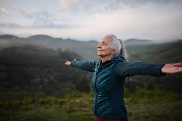 Vanhempi nainen tekee hengitys liikuntaa luonnossa varhain aamulla sumu ja vuoret taustalla. - Valokuva, kuva