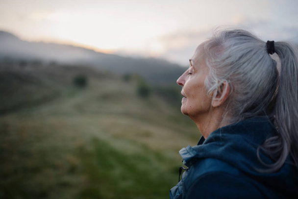 A senior woman doing breathing exercise in nature on early morning with fog and mountains in background. - Valokuva, kuva