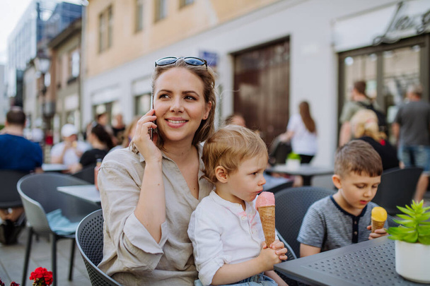 A young mother taking selfie with her kids eating ice-cream in cafe outdoors in street in summer. - Photo, Image