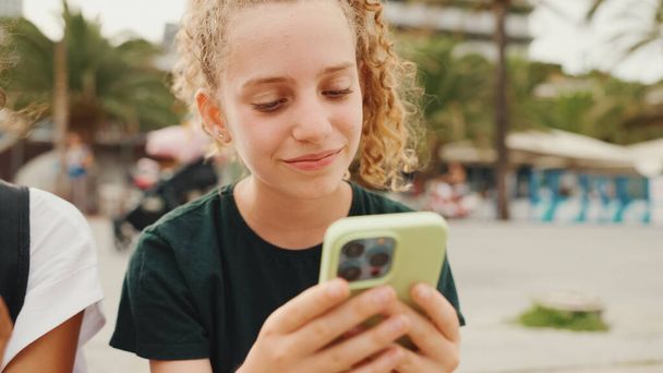 Pre-teenage girl uses mobile phone while sitting on the waterfront. Teenage girl making video call on smartphone - Zdjęcie, obraz