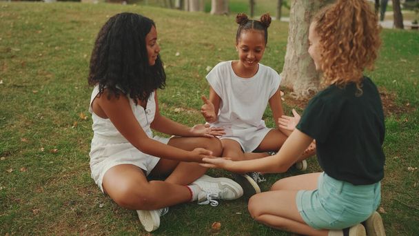 Laughing three girls friends pre-teenage sitting on the grass in the park playing holding each other's hands and clapping their hands - Fotó, kép