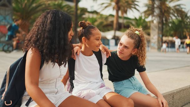 Three girls friends pre-teenage are sitting on the waterfront smiling and emotionally talking holding each other embracing. Teenagers on the outdoors in urban cityscape background - Φωτογραφία, εικόνα