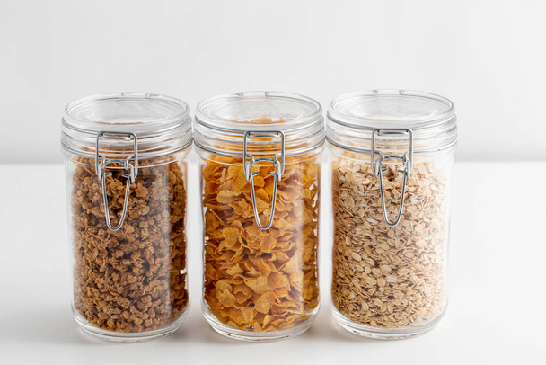 food storage, healthy eating and diet concept - jars with oat, corn flakes and granola on white background - Photo, image