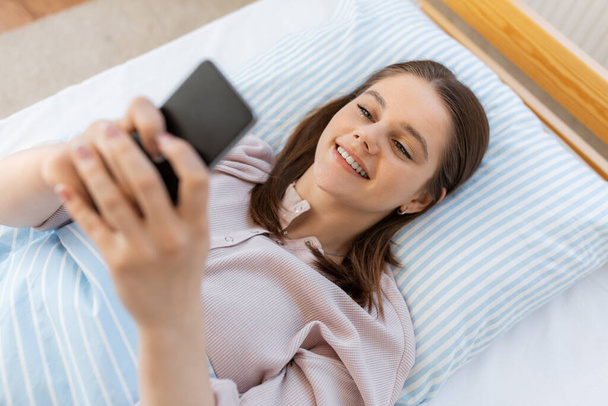 people, bedtime and rest concept - happy smiling teenage girl with smartphone lying in bed at home - Foto, Bild