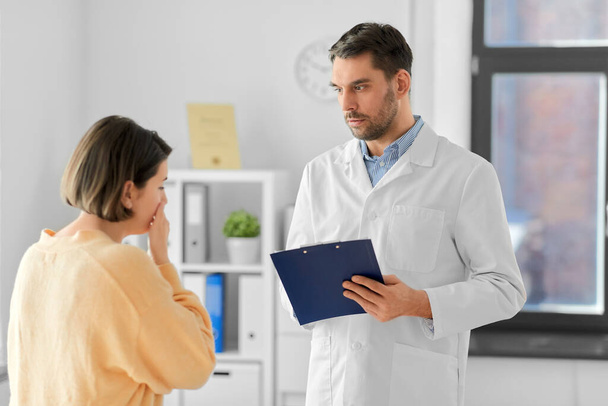 medicine, healthcare and people concept - male doctor with clipboard talking to scared or coughing woman patient at hospital - Foto, Bild
