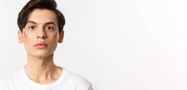 People, lgbtq and beauty concept. Close-up of beautiful androgynous man with glitter on face, looking at camera, standing over white background. - Фото, изображение