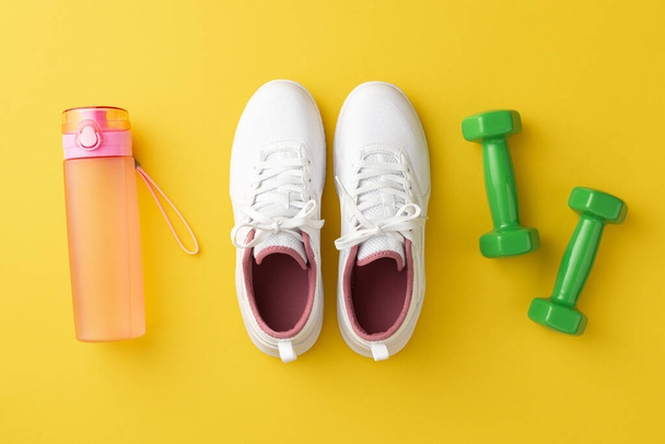 Koncepcja akcesoriów fitness. Top view zdjęcie białe trampki różowa butelka wody i zielone hantle na izolowanym żółtym tle - Zdjęcie, obraz
