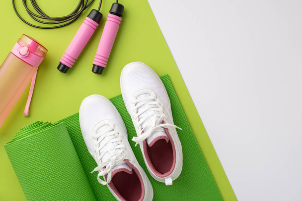 Sports accessories concept. Top view photo of skipping rope pink bottle of water and white sneakers over green exercise mat on bicolor green and white background with copyspace - Fotoğraf, Görsel