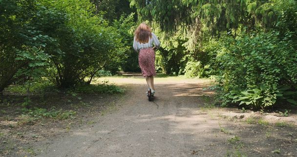 Eine junge Frau fährt mit einem Elektroroller auf einem Weg in einem Park zwischen grünen Bäumen. Rückansicht, tagsüber, Breitbild.. Hochwertiges 4k-Filmmaterial - Foto, Bild