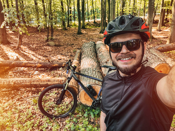 Glücklicher Radfahrer im ländlichen Raum mit seinem Mountainbike im Rücken. Ein lächelnder bärtiger Mann trägt Fahrradhelm und Sonnenbrille, während er im Wald unterwegs ist. Freizeitaktivitäten im Wald. - Foto, Bild