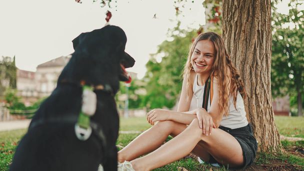 Cute girl with black dog is resting under tree on the lawn. Happy dog rejoices being on walk in the outdoor. Animal training outdoors.  - Valokuva, kuva