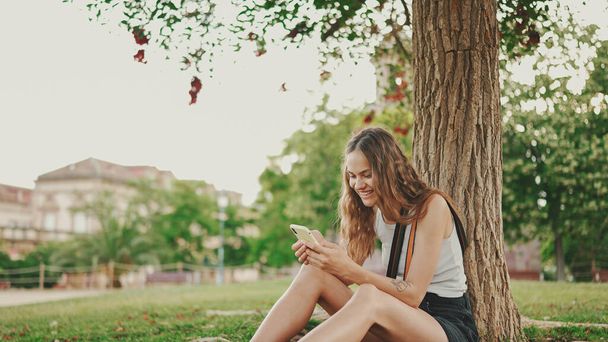 Beautiful girl with long wavy hair wearing in white top uses mobile phone while sitting on the green grass under tree in the park. Girl flip through the pictures on your phone with fingers - Valokuva, kuva
