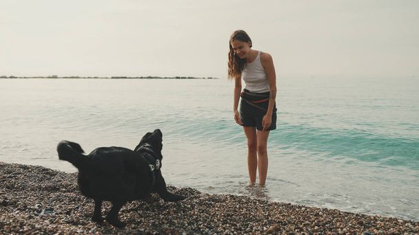 Cute girl with long wavy hair in white top plays on pebble beach with black dog seascape background - Foto, imagen