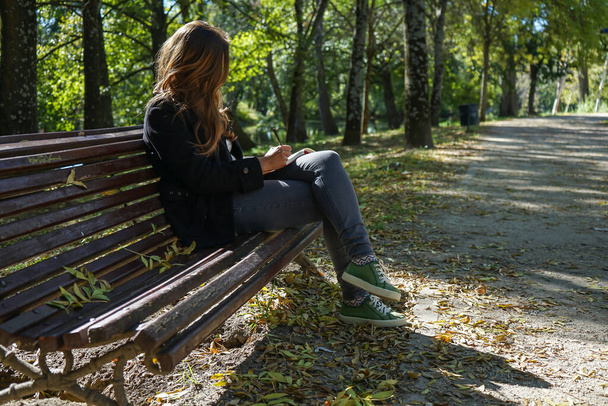 Geconcentreerde vrouw die schrijft of tekent op een notitieboekje zittend op een bankje in een park. - Foto, afbeelding