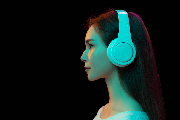 Side view portrait of young girl listening to music in white headphones isolated on black studio background in neon light. Concept of beauty, fashion, emotions, facial expression, youth, lifestyle, ad - Photo, image