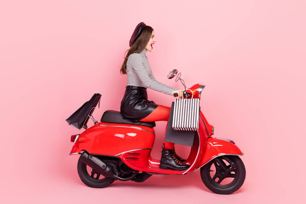 Profiilin puolella kuva tyylikäs trendikäs nainen ratsastaa nopea moottoripyörä ajoneuvon ostoskassit eristetty vaaleanpunainen väri tausta. - Valokuva, kuva