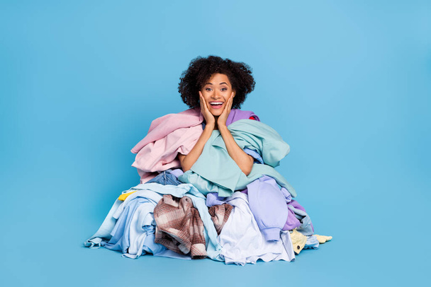 Photo of positive excited woman arms hands cheekbones cheeks covering big clothes stack isolated blue color background. - Fotó, kép