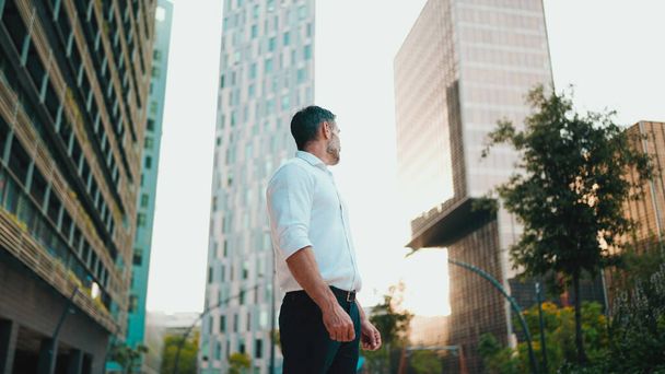 Mature businessman with neat beard wearing white shirt stands in the financial district in the city. Successful man looks around on modern buildings background - Foto, Imagem