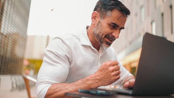 Mature businessman with neat beard wearing white shirt user on laptop pc computer sit at cafe outdoors. Successful man sitting at cafe table outdoors on nature Mobile office freelance - Фото, изображение