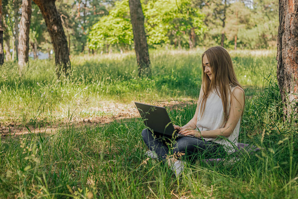 On the banner, a young girl works with a laptop in the fresh air in the park, sitting on the lawn. The concept of remote work. Work as a freelancer. The girl takes courses on a laptop and smiles - Foto, immagini