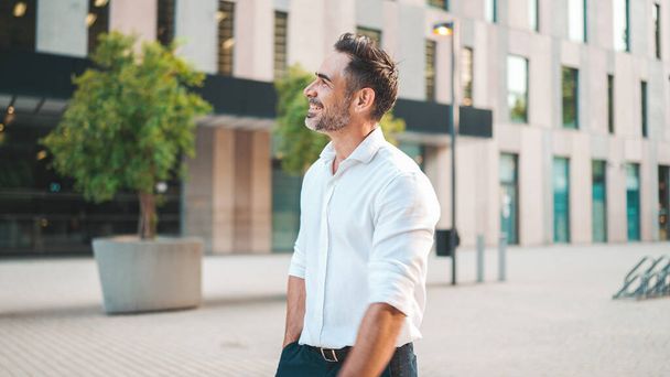 Mature businessman with neat beard wearing white shirt on his way to the office in the financial district in the city. Successful man Looks at the upper floors of modern buildings - Foto, Bild