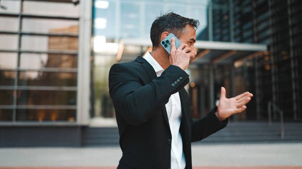 Middle-aged businessman with neat beard in office clothes uses mobile phone. Successful man emotionally talking on smartphone. Disappointment, trouble - Photo, image