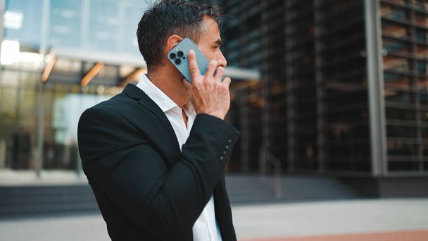 Middle-aged businessman with neat beard in office clothes uses mobile phone. Successful man emotionally talking on smartphone. Disappointment, trouble - 写真・画像
