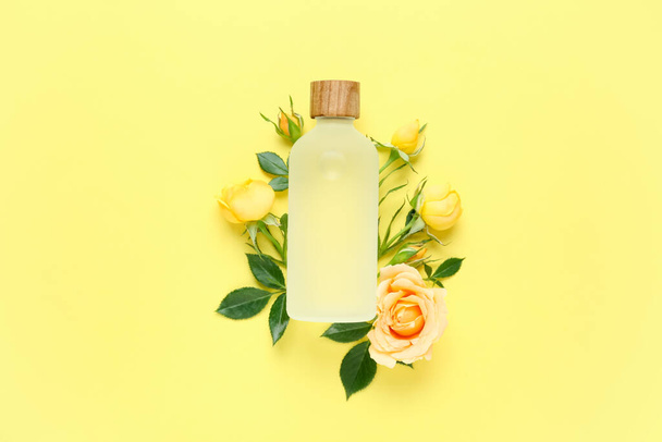 Bottle of rose essential oil and beautiful flowers on pale yellow background, top view - Фото, изображение