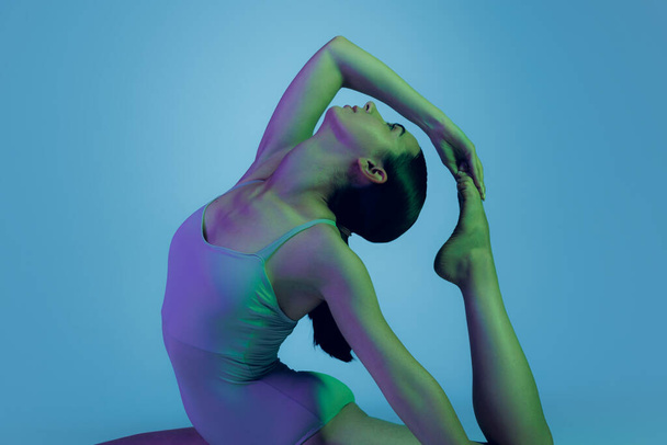 Portrait of young sportive girl doing stretching exercises isolated over blue studio background in neon light. Flexible body. Concept of sport, health, hobby, youth, strength, lifestyle, ad - Foto, Imagem