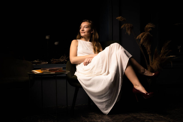 Egy fehér ruhás fiatal nő üldögél egy karosszéken egy sötét szobában.. - Fotó, kép