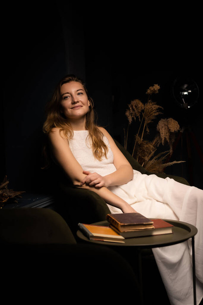A young woman in a white dress smiles and sits relaxed on an armchair in a dark room. - Foto, immagini
