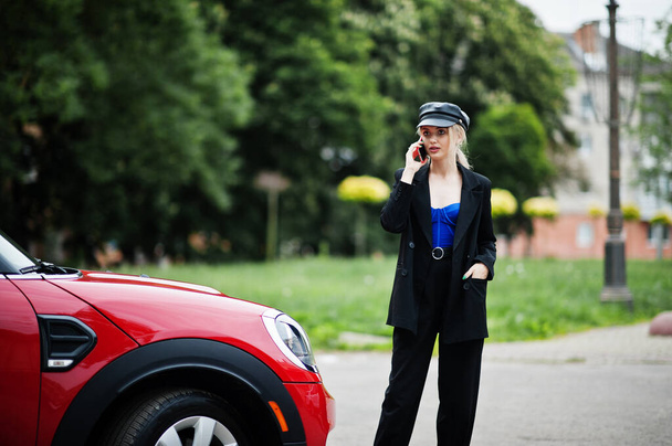 Portrait of beautiful blonde sexy fashion woman model in cap and in all black with bright makeup near red city car, with mobile phone. - Фото, зображення