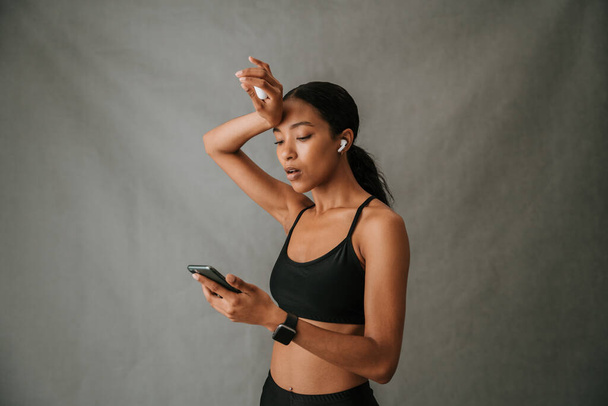 Мультиэтничная женщина, озадаченная сотовым телефоном с помощью наушников после тренировки. Высокое качество фото - Фото, изображение