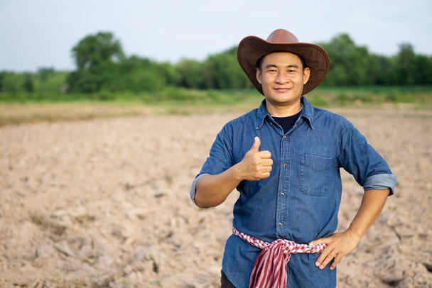 Przystojny Azjata jest na plantacji. Nosi kapelusz, niebieską koszulę, tajskie przepaski na talii, kładzie rękę na biodrach, kciuki w górę, czuje się pewnie, patrzy w kamerę. Pojęcie: Zawód rolnika.  - Zdjęcie, obraz