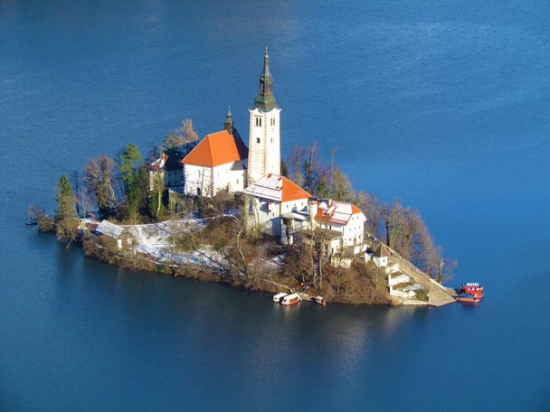 Vista de una iglesia en la isla en el lago Bled en la región de Gorenjska de Eslovenia con barcos ancored en las escaleras que conducen a la parte superior - Foto, Imagen