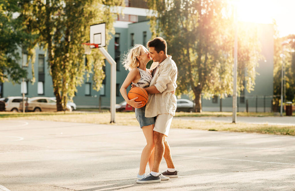 sport, love and people concept - happy young couple with ball hugging on basketball playground - Fotó, kép