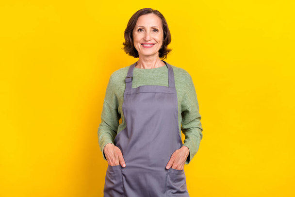 Portrait of attractive cheerful grey-haired woman barista helper holding hands in pockets isolated over bright yellow color background. - Fotó, kép