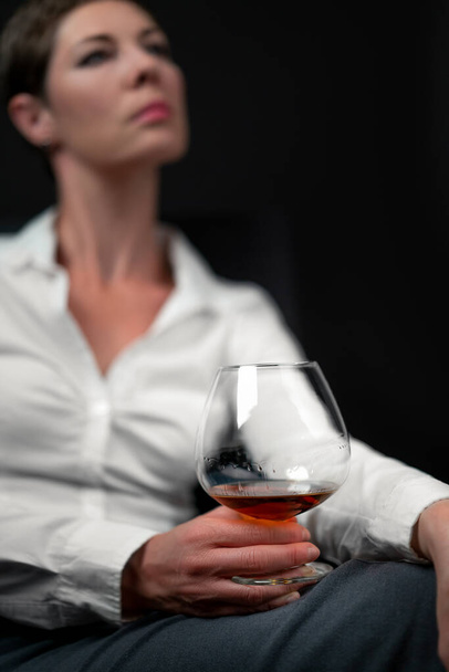 Drinking glass filled with alcohol held by sad looking woman in business clothes sitting with her legs crossed. Womans image blurred. Concept for depression, sadness, alcoholism, drinking problems - Фото, зображення