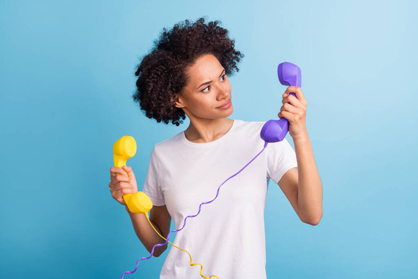 Portrait of young funky funny smiling joyful pretty afro girl hold two telephones doubtful isolated on blue color background. - Foto, immagini