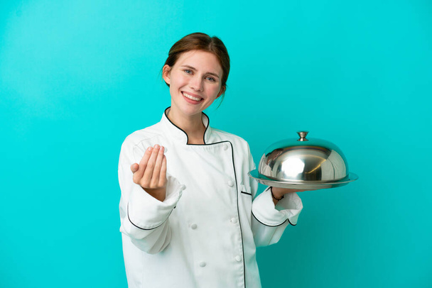 Young chef woman with tray isolated on blue background inviting to come with hand. Happy that you came - 写真・画像