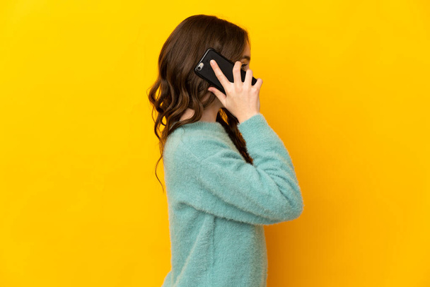 Little caucasian girl isolated on yellow background keeping a conversation with the mobile phone with someone - Fotó, kép