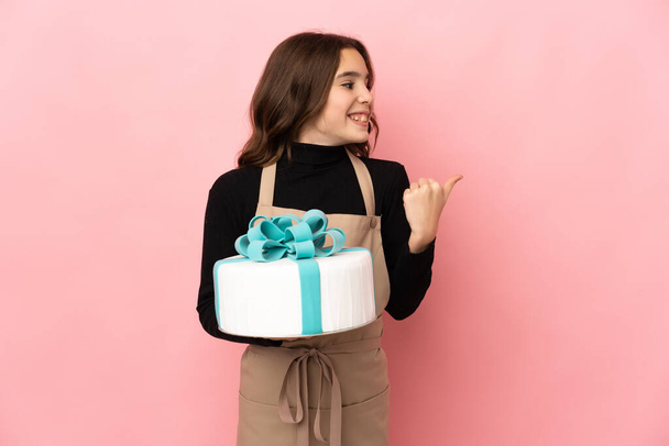 Little Pastry chef holding a big cake isolated on pink background pointing to the side to present a product - Foto, afbeelding