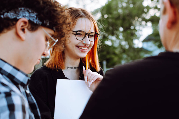 Portrait of young smiling happy woman student with ginger hair wearing black shirt, glasses, holding orange pen, blank sheet of paper, talking to friends young men students. Education, youth, exams. - Photo, image