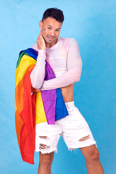 chico gay sonriendo con la bandera del orgullo sobre su hombro, fondo azul. Foto de alta calidad - Foto, Imagen