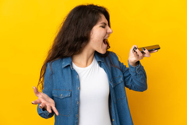 Teenager Russian girl isolated on yellow background using mobile phone and singing - Valokuva, kuva
